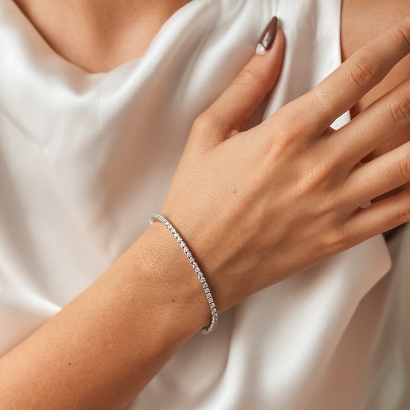 A close-up of a wrist wearing a silver tennis bracelet adorned with zirconia stones, contrasted against a white satin fabric.