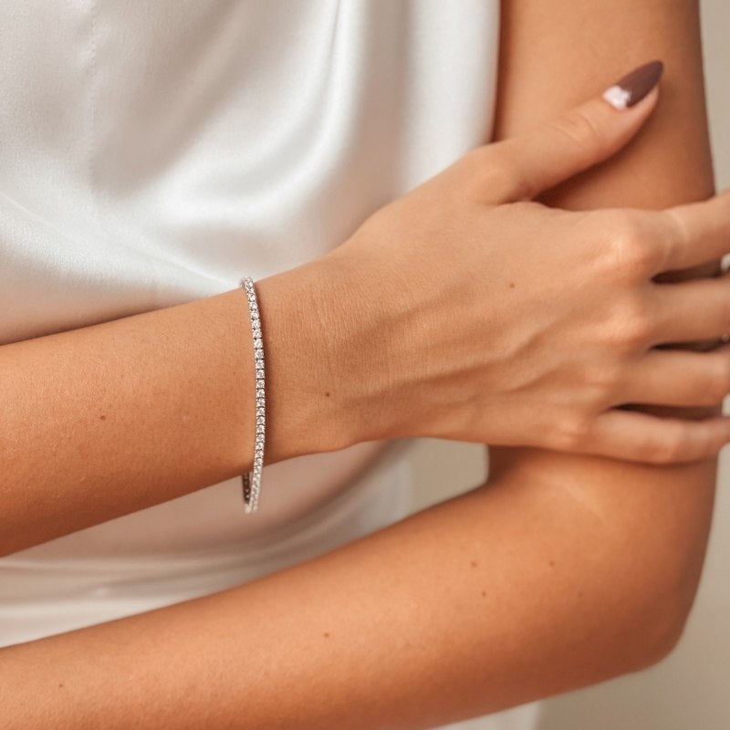 Close-up of a woman's arm wearing a silver tennis bracelet with zirconia stones, against a white satin background.