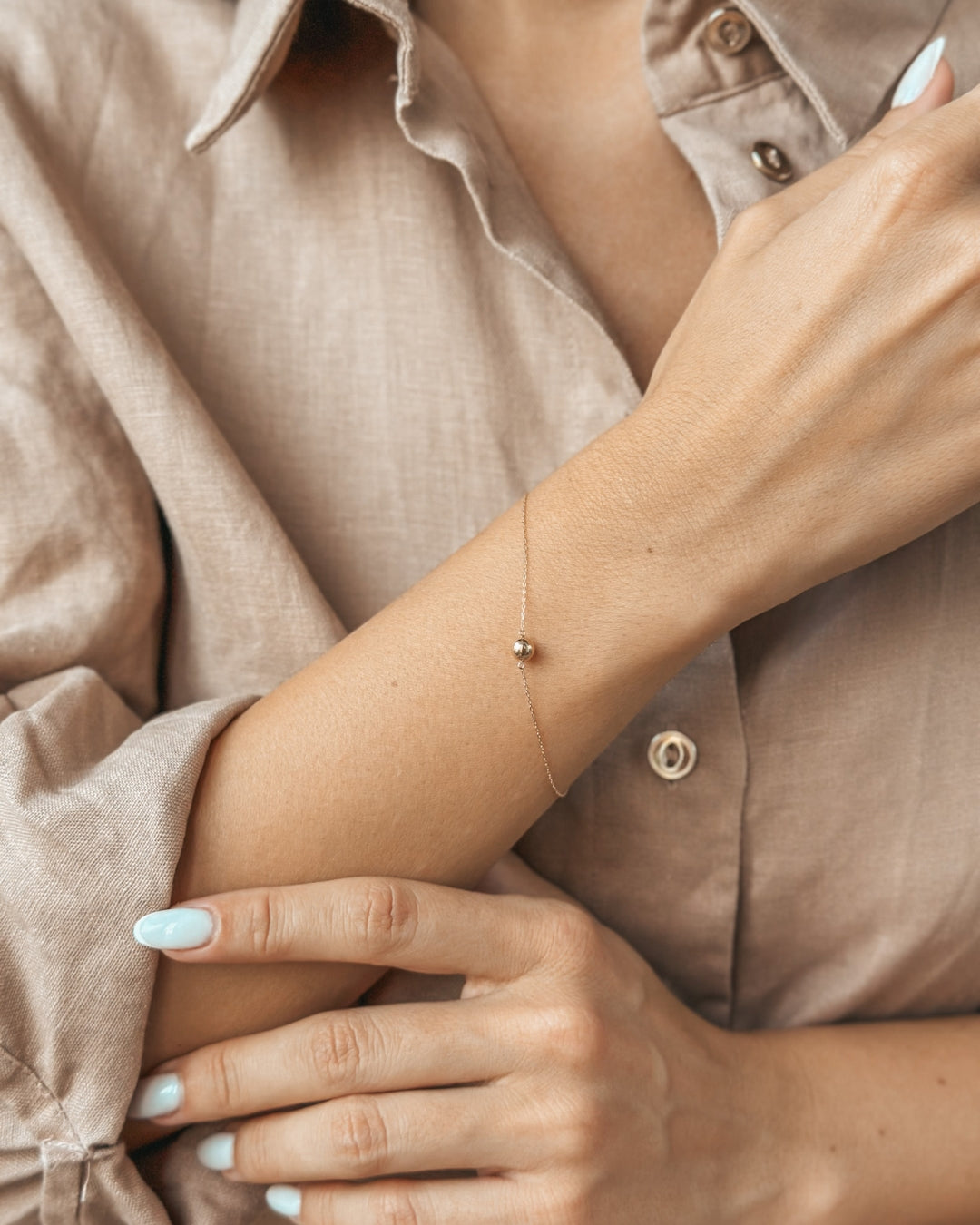Close-up of a person wearing a delicate gold bracelet on their wrist, with a beige shirt sleeve helping accentuate the elegance of the accessory.
