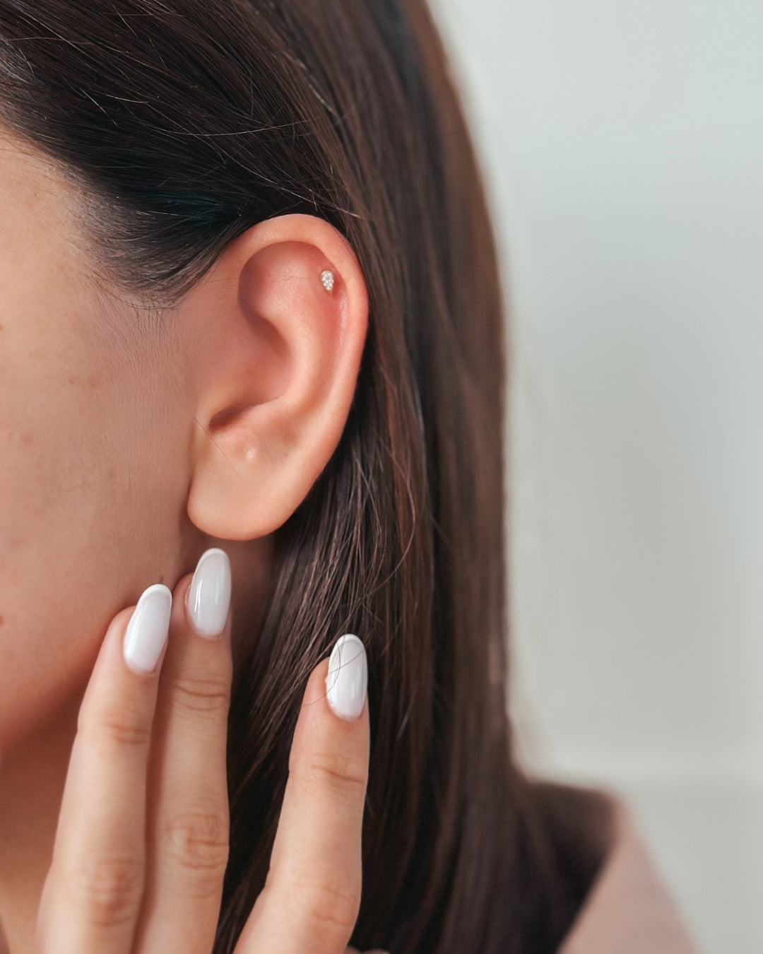 Close-up of an ear with a small stud earring and fingers with white polished nails.