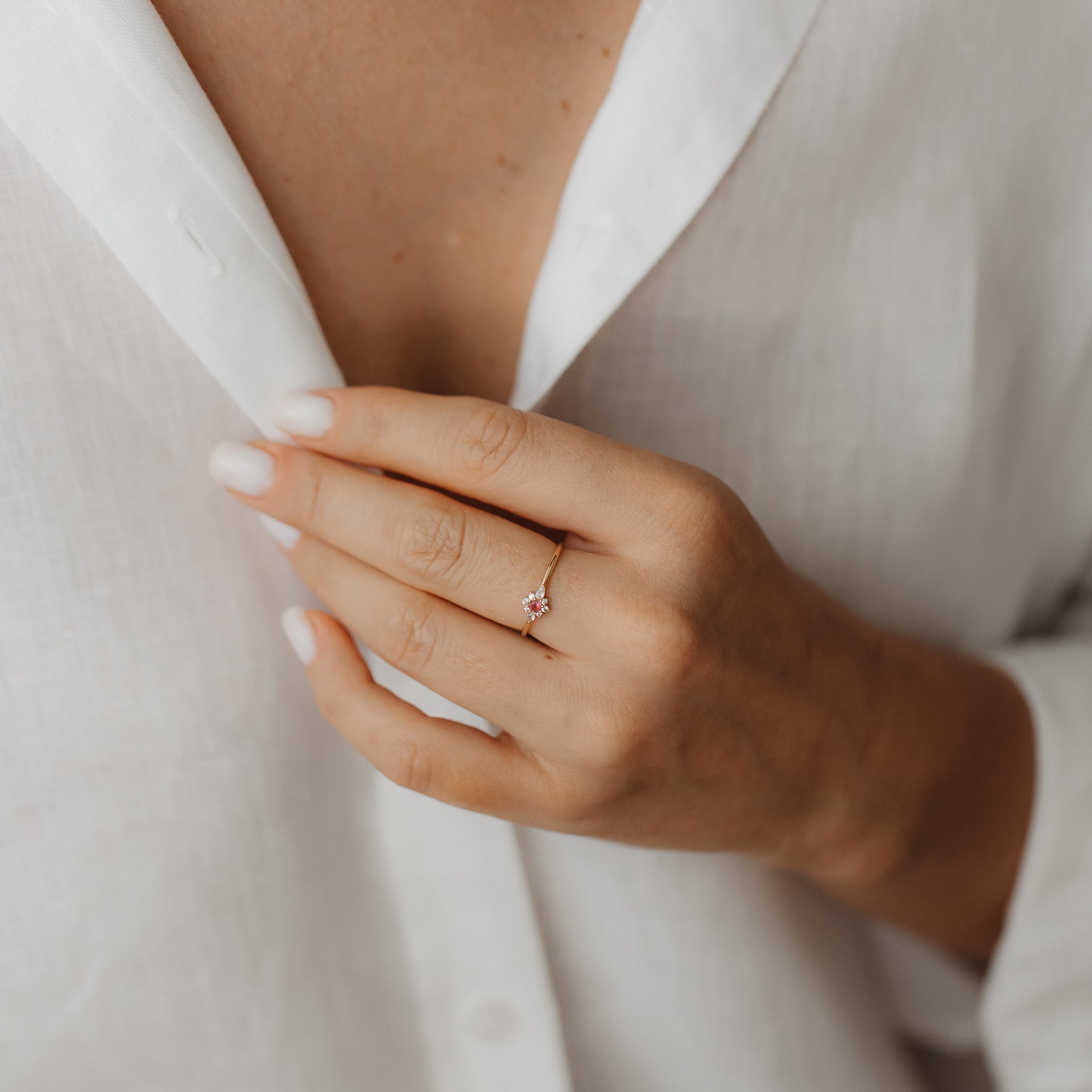 Close-up of a hand wearing the Flora 14K yellow gold ring with a white sapphire and pink tourmaline floral cluster, against a white linen shirt.
