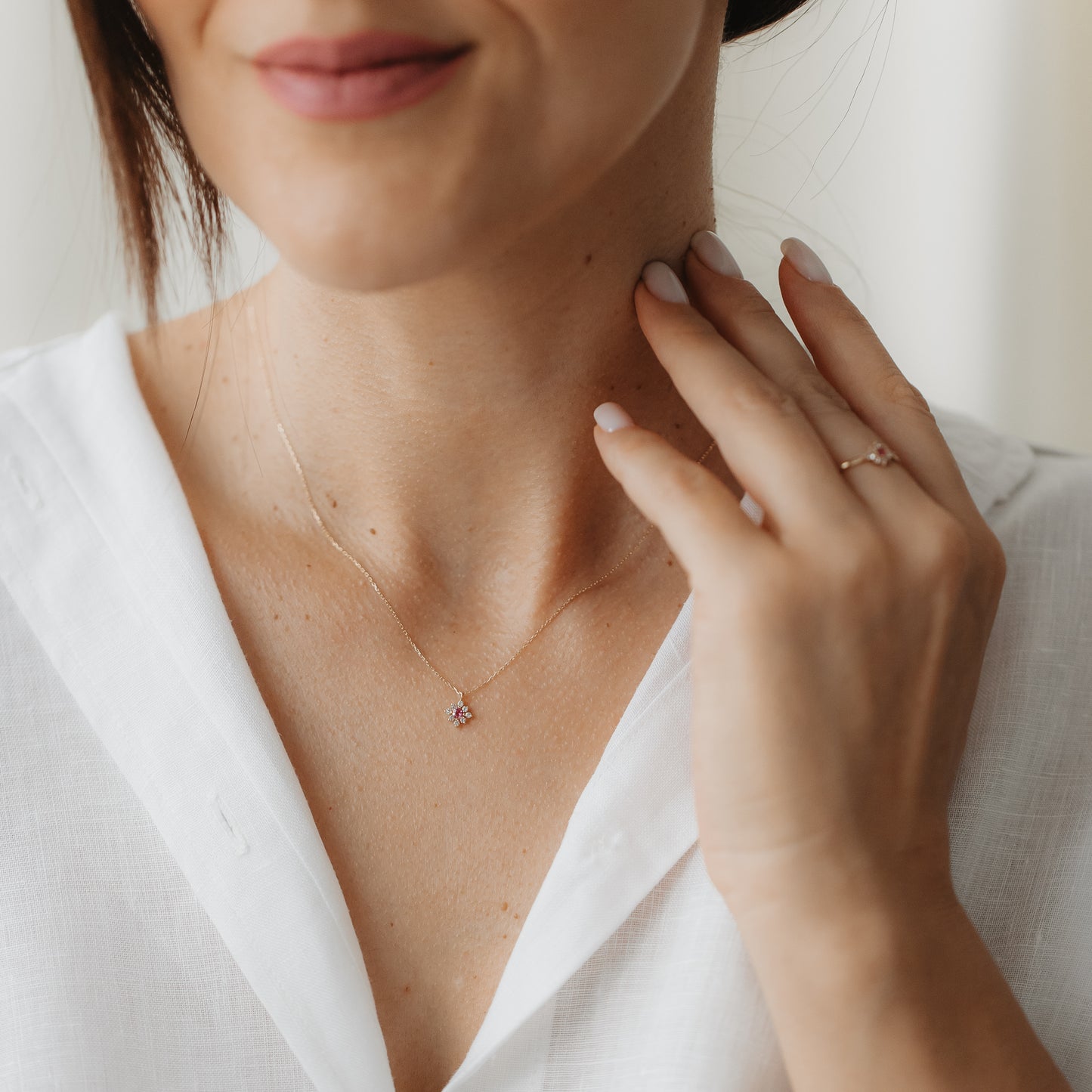 Close-up of a person wearing the Flora 14k gold necklace with a tiny floral flower pendant set with pink tourmaline and white sapphires, hand touching the neck while wearing a white blouse.