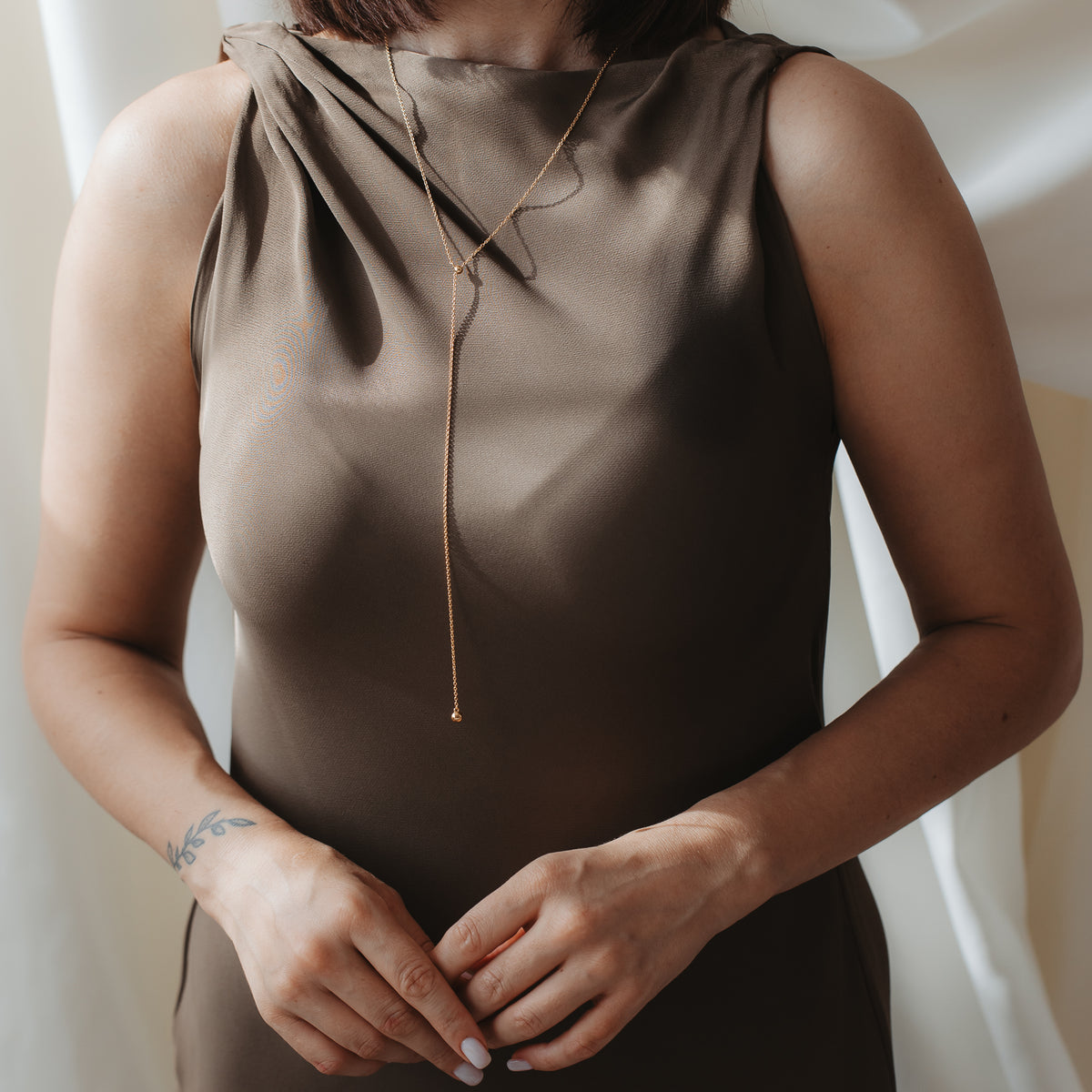 Close-up of a person wearing a gold‑plated 925 silver bola lariat necklace over a brown satin dress, hands gently clasped.