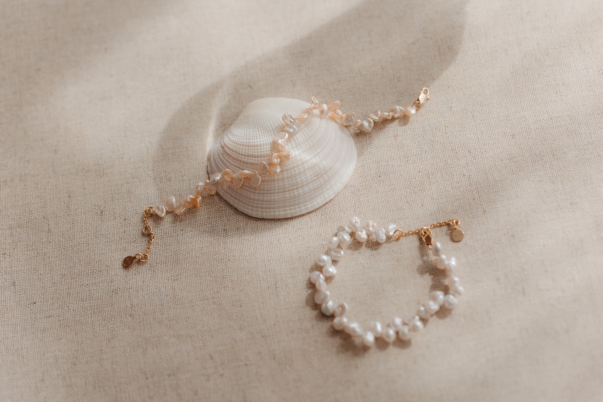 Two gold‑plated sterling silver bracelets with pink keshi pearls styled on a seashell over beige linen fabric.