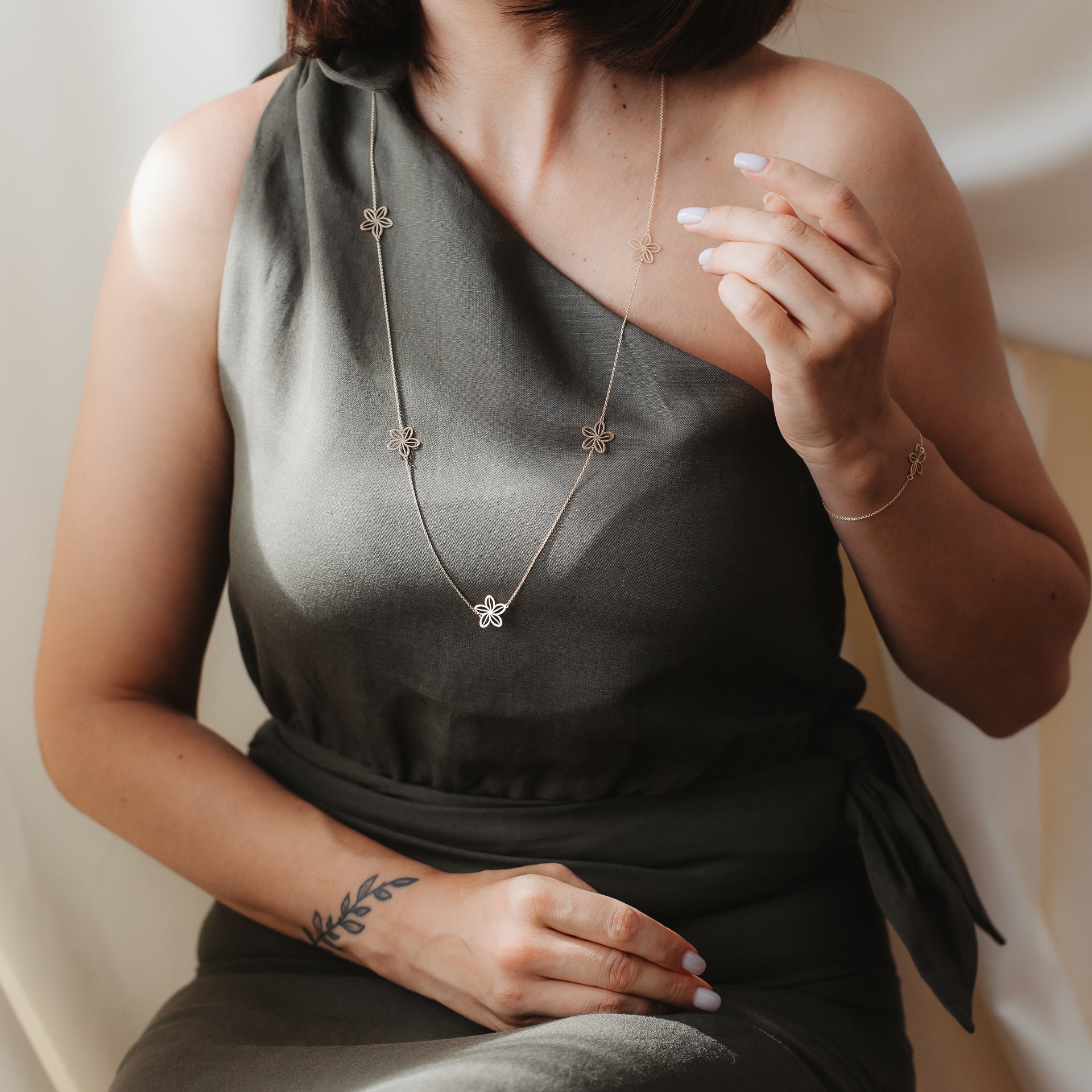 Közeli felvétel egy olívazöld, egyvállas ruhát viselő modellről, aki a Wildflower Maxi 14K yellow gold station necklace nyakláncot visel apró virágmedálokkal, lágy beltéri fényben, hozzá illő karkötővel.
