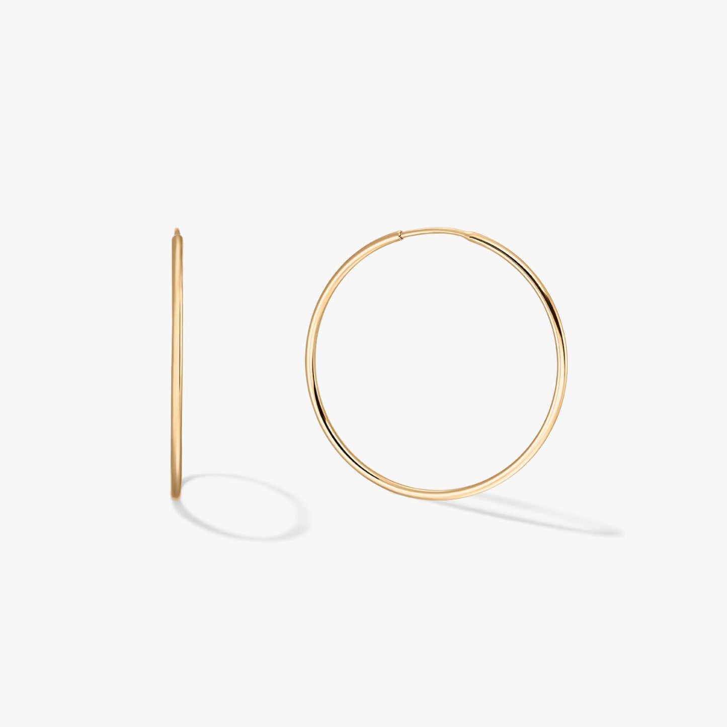 Pair of large thin 14K yellow gold hoop earrings on a white background, one shown front-on and one in profile with soft shadows.