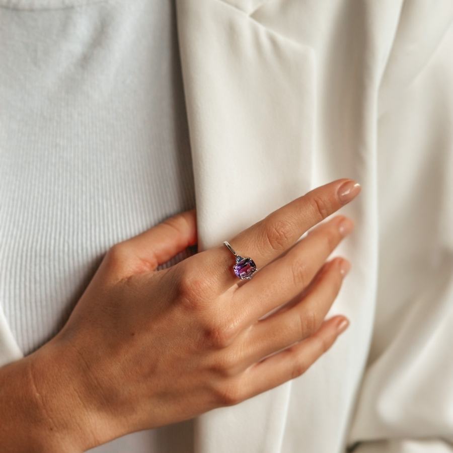 Close-up of a hand wearing a silver ring with amethyst and tanzanite gemstones, resting on a white garment.