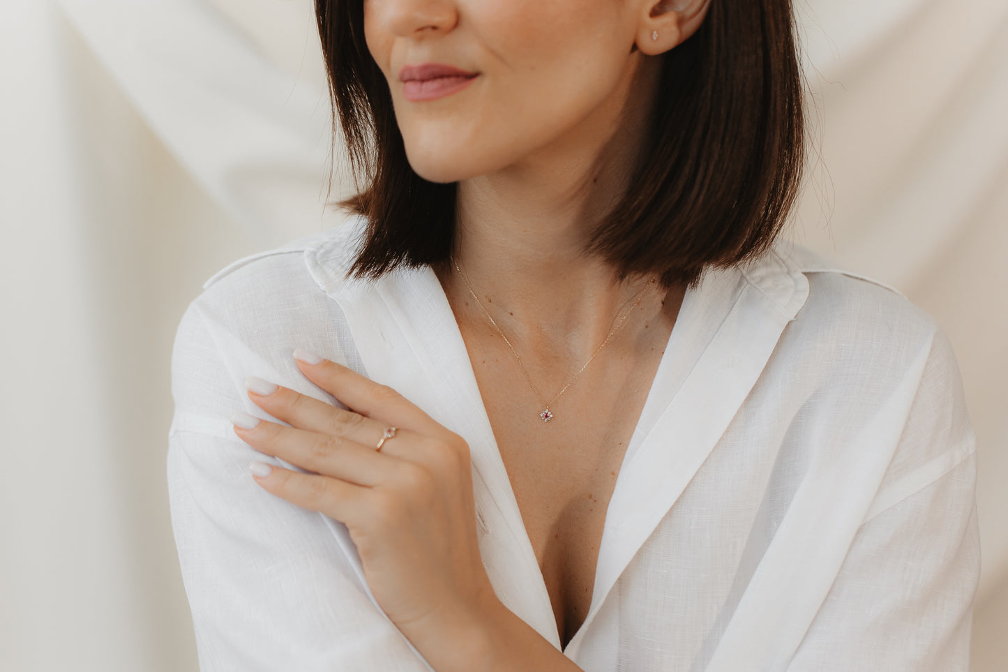 Close-up of a person in a white blouse wearing a delicate 14K yellow gold ring with a pink tourmaline and white sapphire, hand resting on the shoulder against a soft neutral background.
