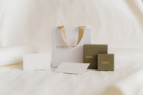 Minimal studio photo of INDIRA gift packaging set: a white bag with gold ribbon handles, two olive-green jewelry boxes, and blank cards on a soft ivory background.