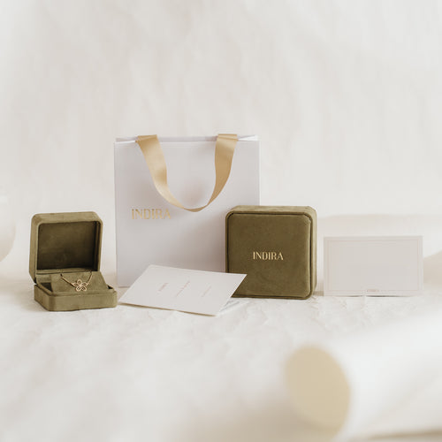 Indira branded gift packaging set with a white bag and gold ribbon handles, olive velvet jewelry boxes (one open), and cards arranged on a soft neutral background.
