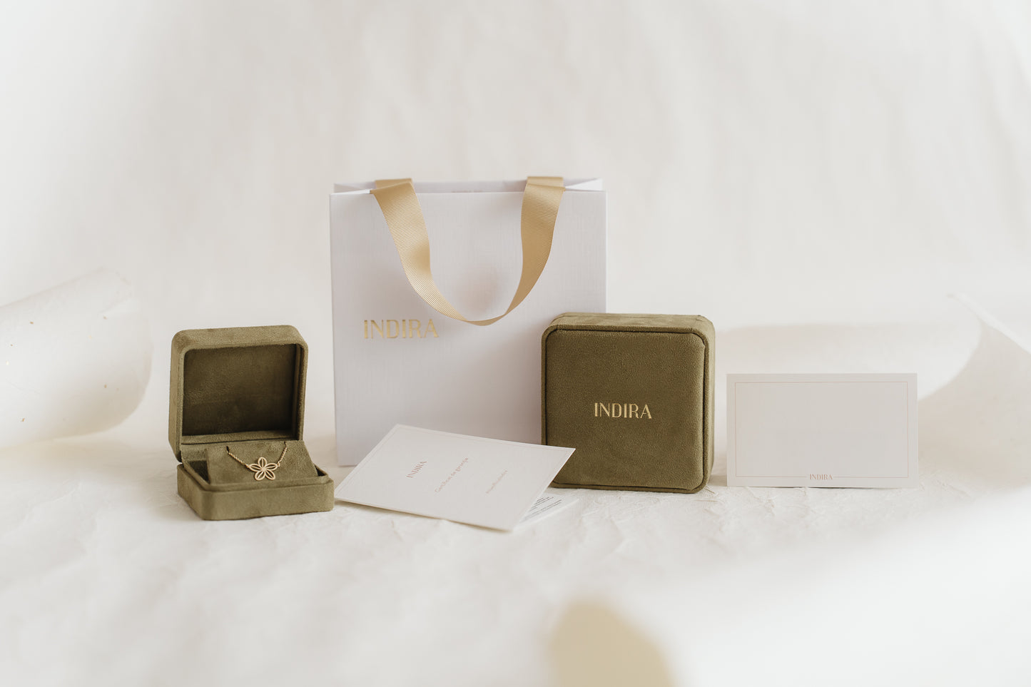 Indira gift packaging set: white shopping bag with gold ribbon handles, two olive velvet jewelry boxes (one open showing a gold flower pendant), and branded cards on a soft white studio background.