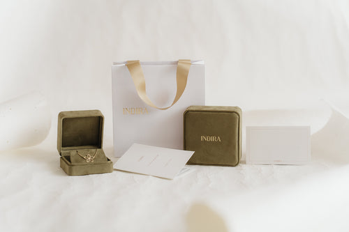 Indira gift packaging set: white shopping bag with gold ribbon handles, two olive velvet jewelry boxes (one open showing a gold flower pendant), and branded cards on a soft white studio background.