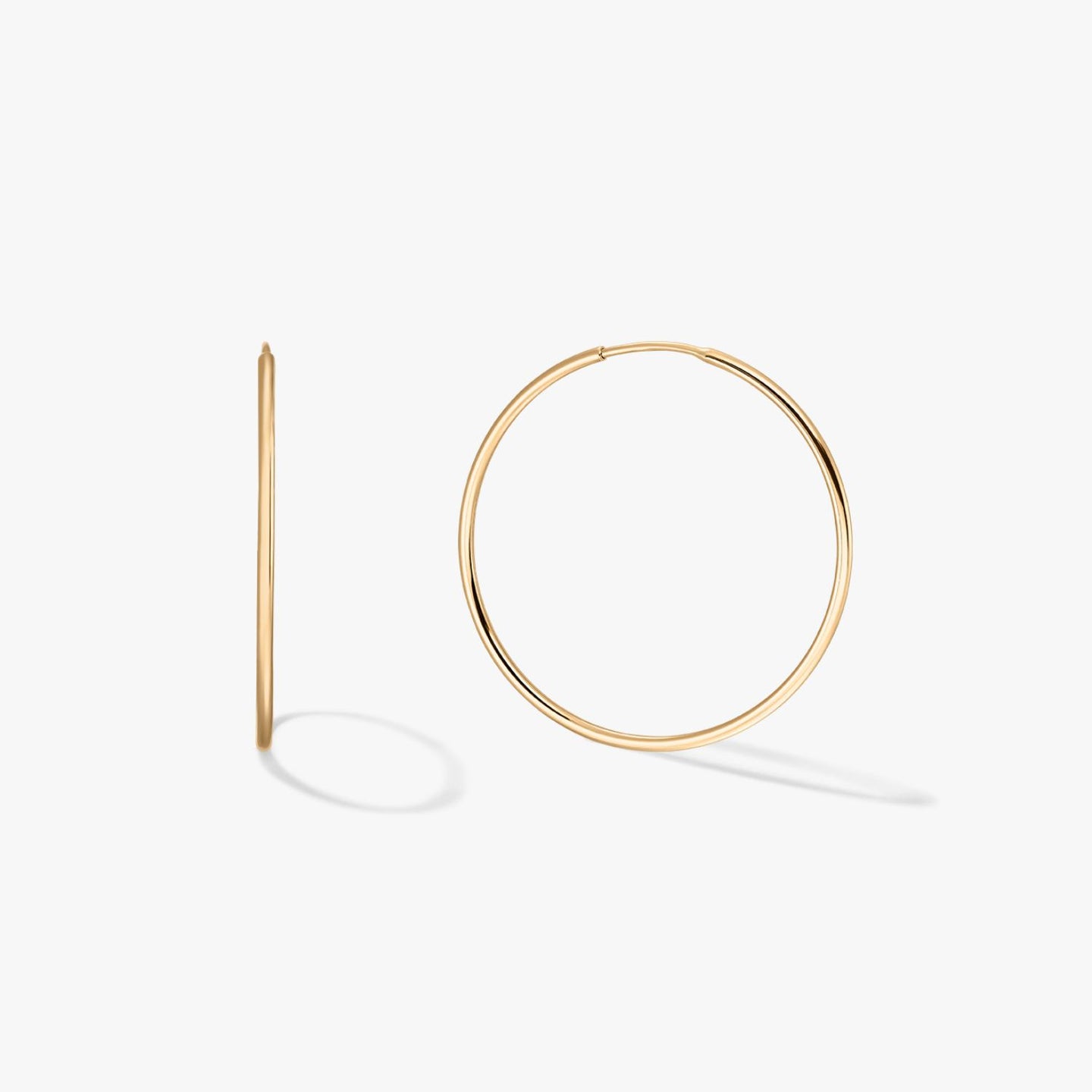 Pair of large thin 14K yellow gold hoop earrings on a white background, one shown front-on and one in profile with soft shadows.