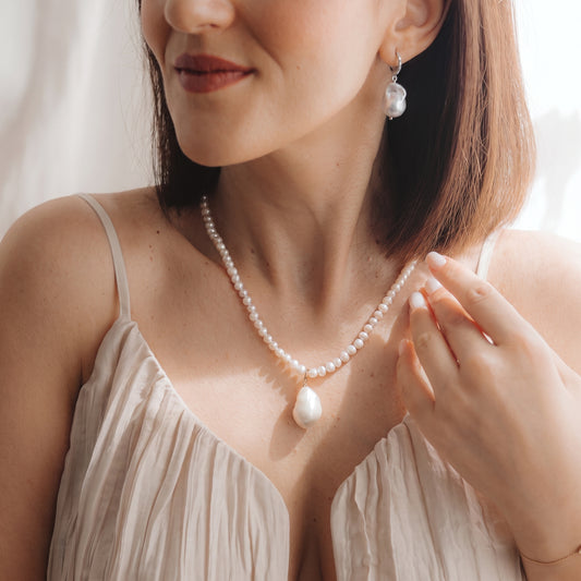 Close-up of a model wearing a rhodium-plated 925 silver baroque pearl drop earring with a small zirconia detail and a matching pearl necklace, hand near the collarbone in soft natural light.