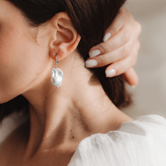 Close-up of an ear wearing a rhodium‑plated 925 silver hoop with a large baroque pearl drop earring, with a hand touching the hair.